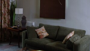 Un salon avec un canapé vert, deux coussins décoratifs, une table basse en verre avec un bol, une table d’appoint avec une lampe et une grande œuvre d’art encadrée au mur.