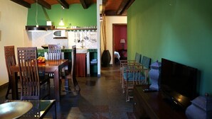 Un coin repas avec des chaises et une table en bois, une cuisine aux murs verts et aux poutres apparentes, et un espace de vie avec un téléviseur et des fauteuils.