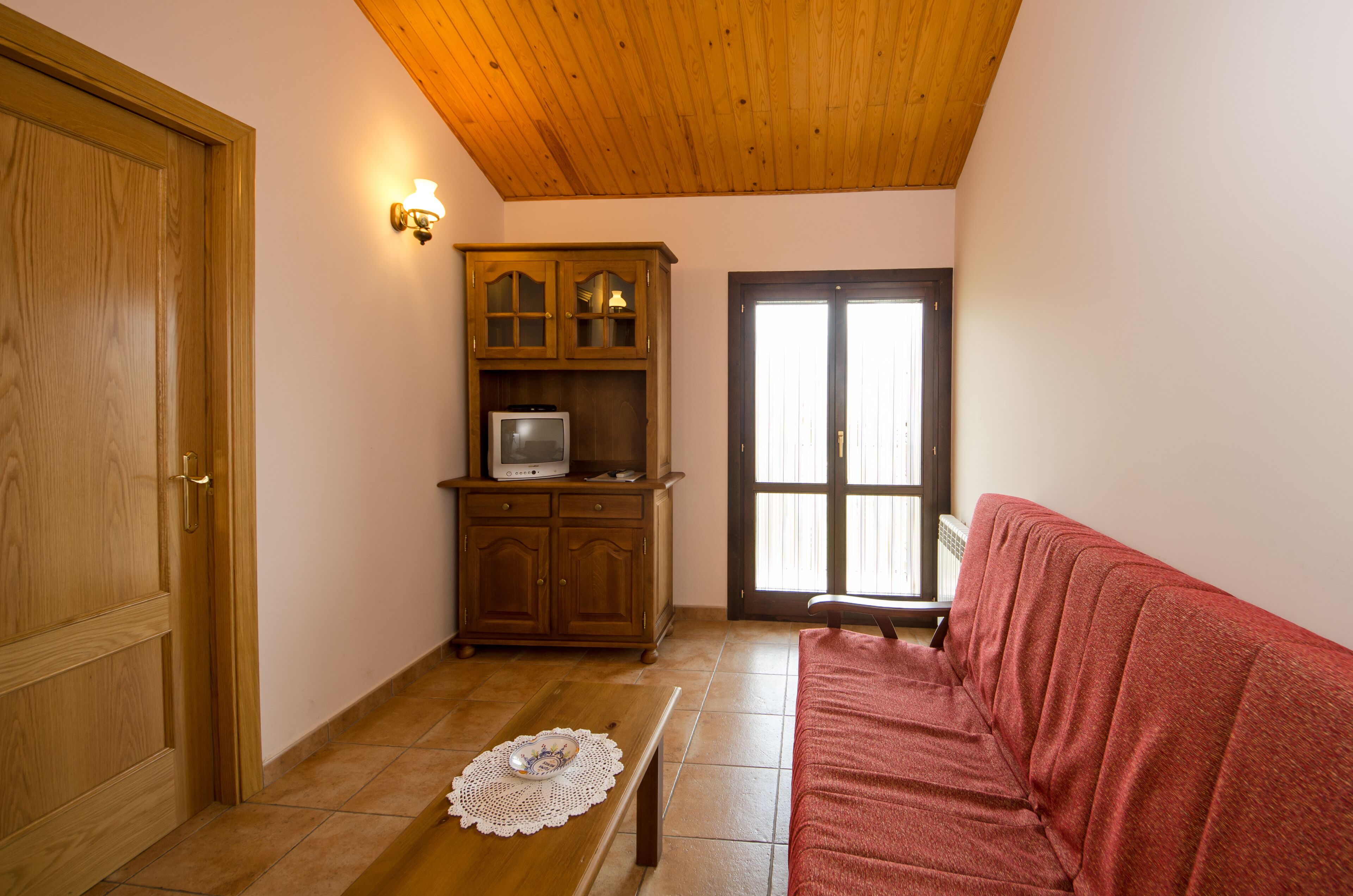 A room with a wooden ceiling, a wooden cabinet with a television, a red sofa, and a door.