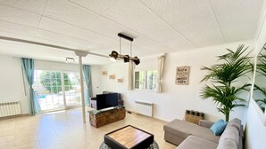 A spacious living room with a sofa, a coffee table, and a TV on a wooden stand. There are large windows with curtains, a plant, and a sign on the wall.