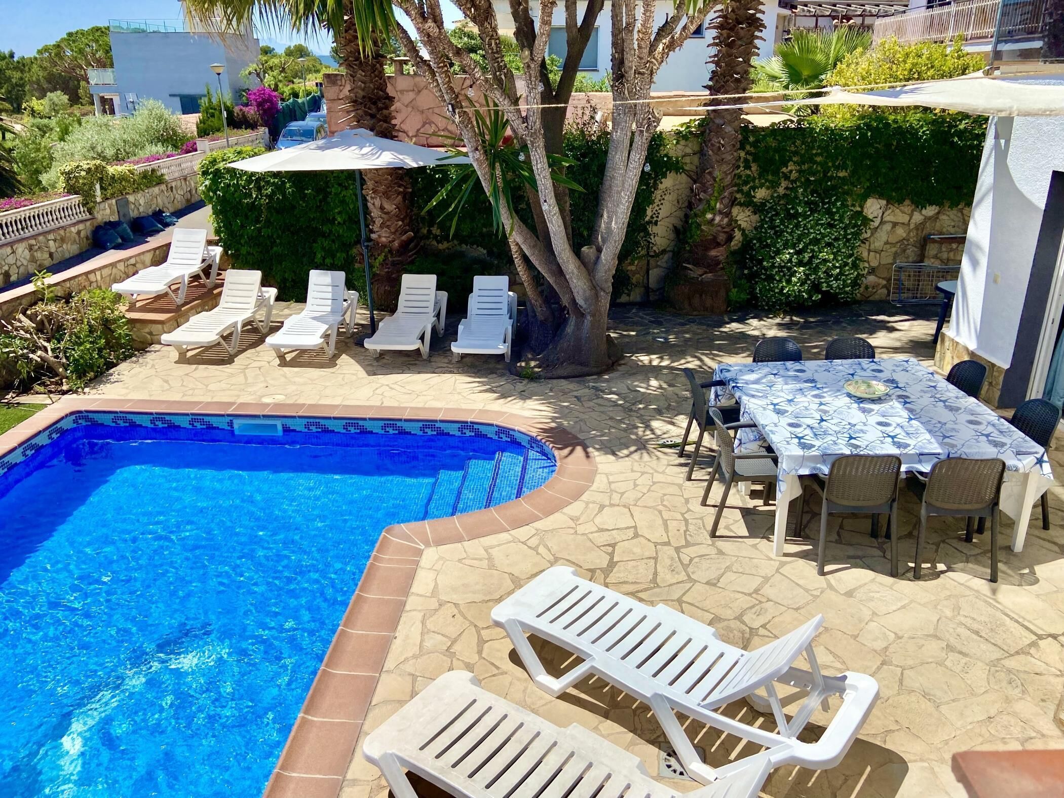 A swimming pool with white lounge chairs and a table with chairs, surrounded by greenery and a stone wall.