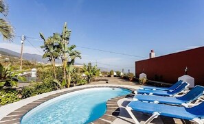Una piscina con sillas de descanso, rodeada de vegetación y un edificio rojo.