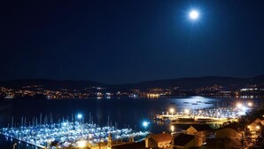 Ein Yachthafen bei Nacht mit vertäuten Booten, ein auffälliger Mond am Himmel und Stadtlichter im Hintergrund.