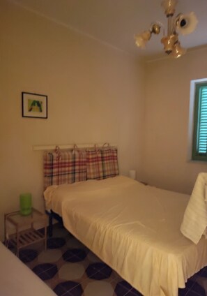 Ein Schlafzimmer mit einem Bett, einem kleinen Nachttisch, einer grünen Mülleimer, einem gemusterten Teppich und einer Deckenleuchte.