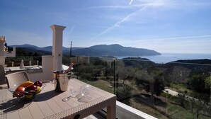 Un balcone con un tavolo, una ciotola di frutta e un bicchiere di vino. Si vede un'area d'acqua e montagne in lontananza.