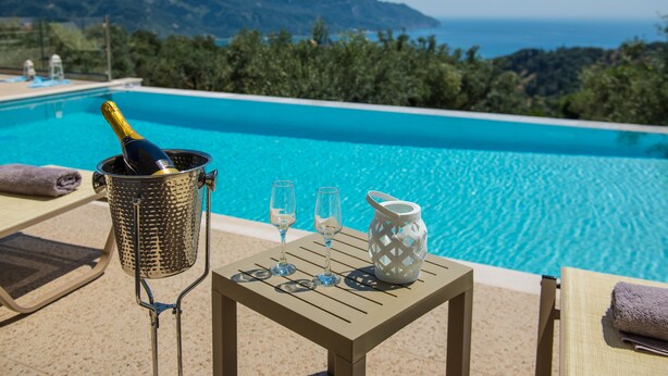 Area piscina con una bottiglia di champagne, due calici e una brocca su un tavolo, con vista su una piscina azzurra e un paesaggio montuoso.
