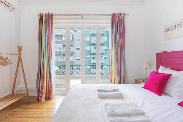 Habitación de hotel con una cama, perchero de madera, balcón con cortinas y vistas a edificios.