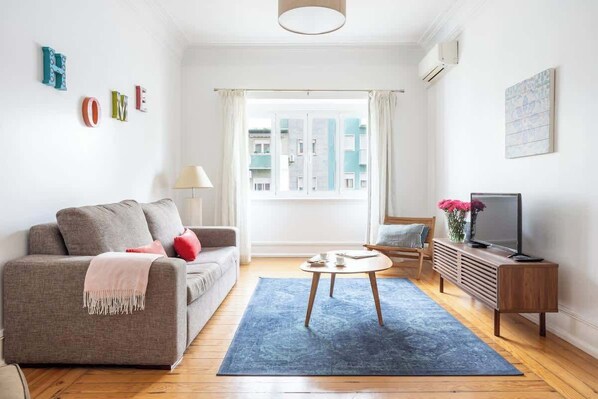 A living room with a sofa, a coffee table, a TV stand, and a window with curtains.