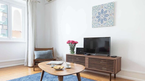 Un salon avec une table basse ronde en bois, un téléviseur posé sur un meuble en bois, un tapis bleu à motifs, un canapé et un élément décoratif mural.