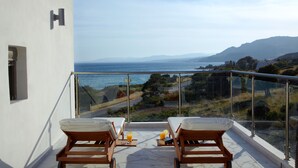 A balcony with two wooden lounge chairs, a small table with a tray of orange juice, and a glass railing overlooking a coastal landscape with mountains in the distance. - Villa Armonia, Exclusive 4 bedroom villa with private heated pool (Lardos)