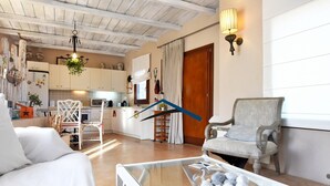 Ein gemütliches Wohnzimmer mit einem Glastisch, einem weißen Sofa und einem grauen Sessel. Im Hintergrund ist eine Küche mit weißen Schränken und einer Holzbalkendecke zu sehen.