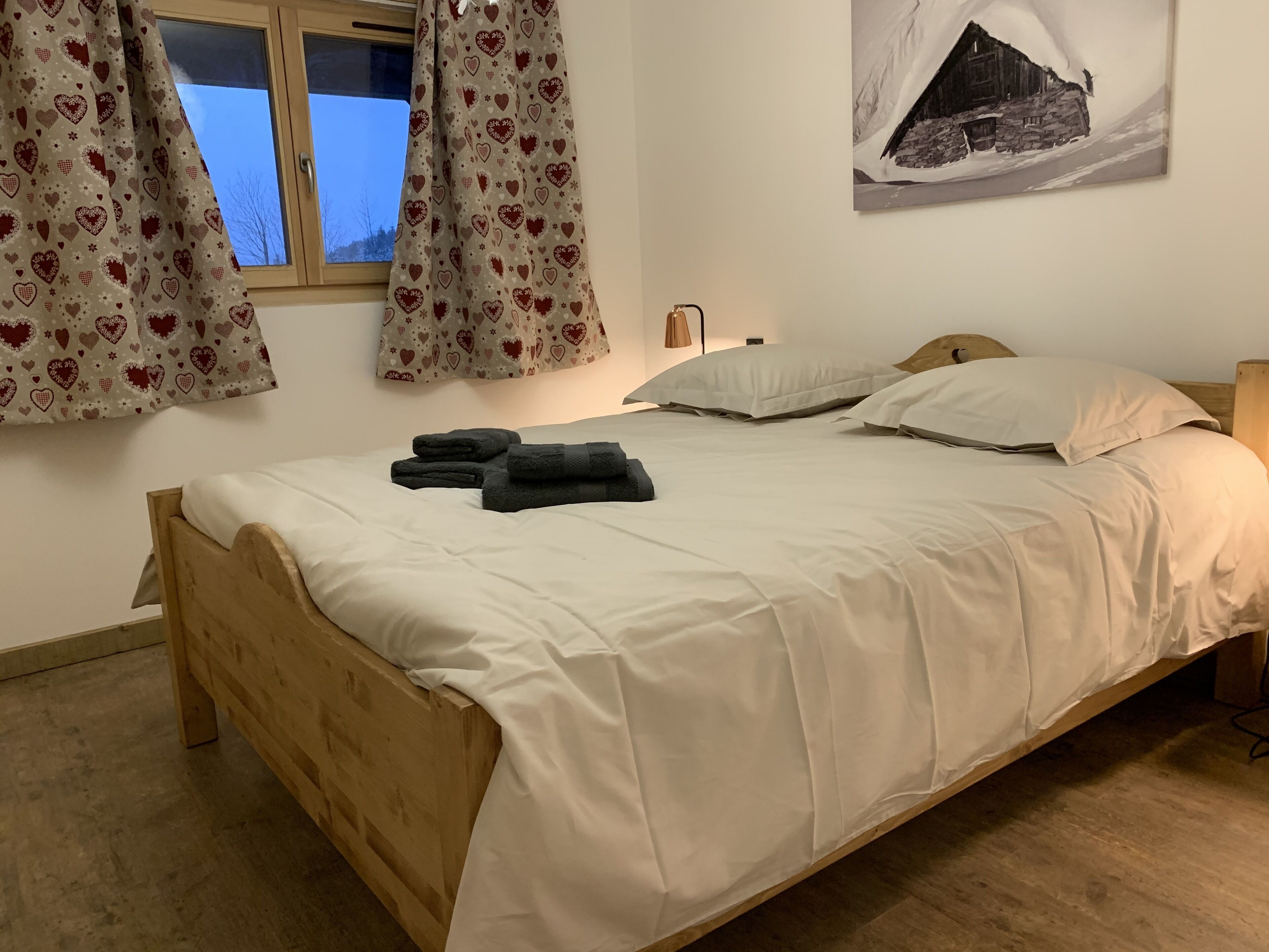 A neatly made bed with white bedding and two pillows, a wooden headboard, and a window with heart-patterned curtains.
