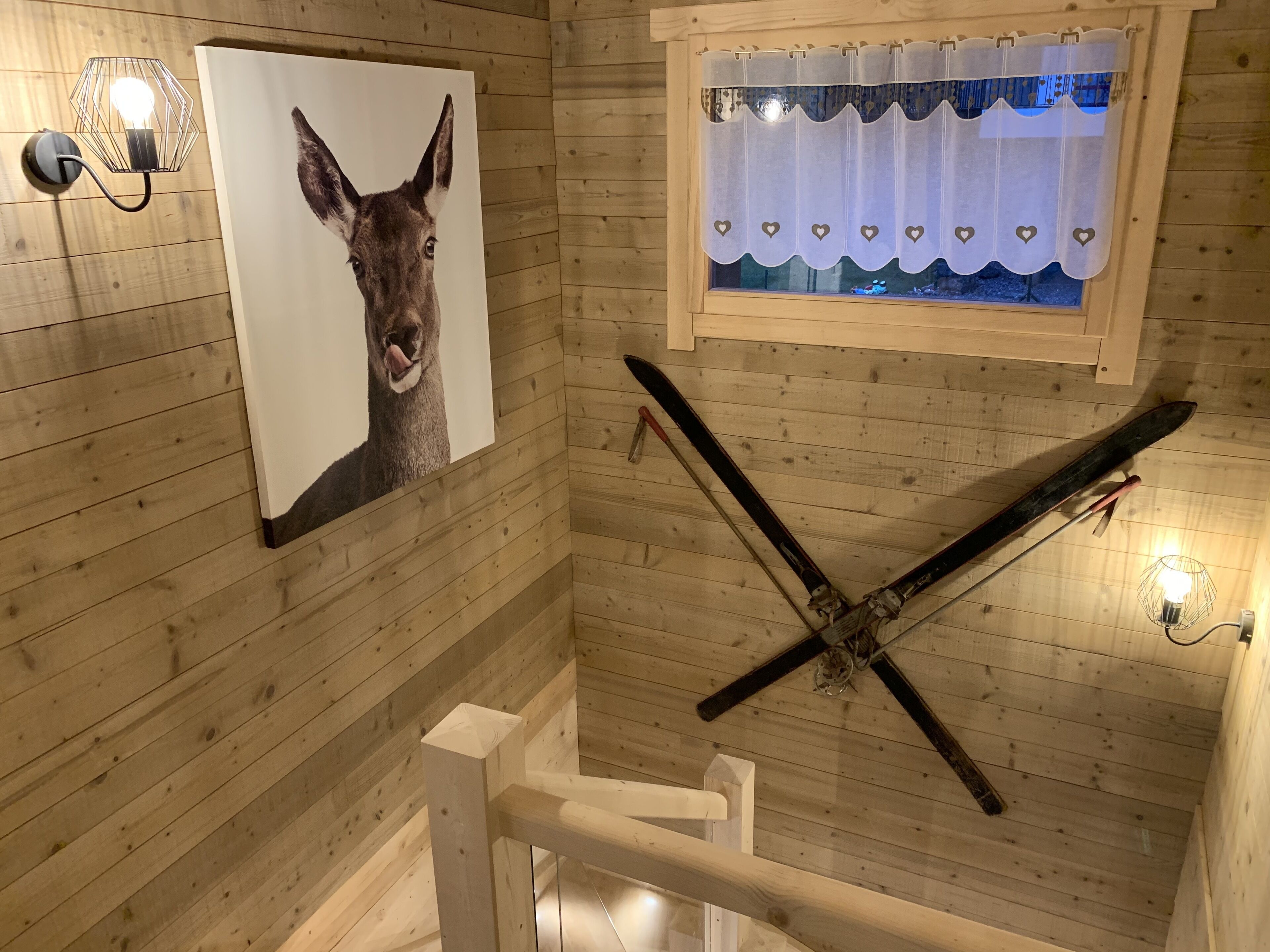 A wooden cabin interior with a ski rack, a wall-mounted light fixture, and a window with a curtain.