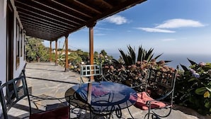 A covered patio with a table and chairs, a view of the ocean, and tropical plants.