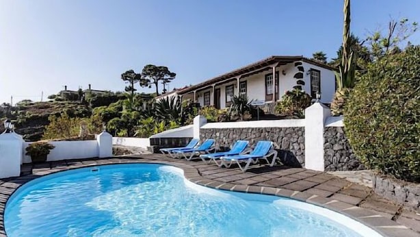 A swimming pool with blue water, surrounded by a stone wall and a white building with a tiled roof.