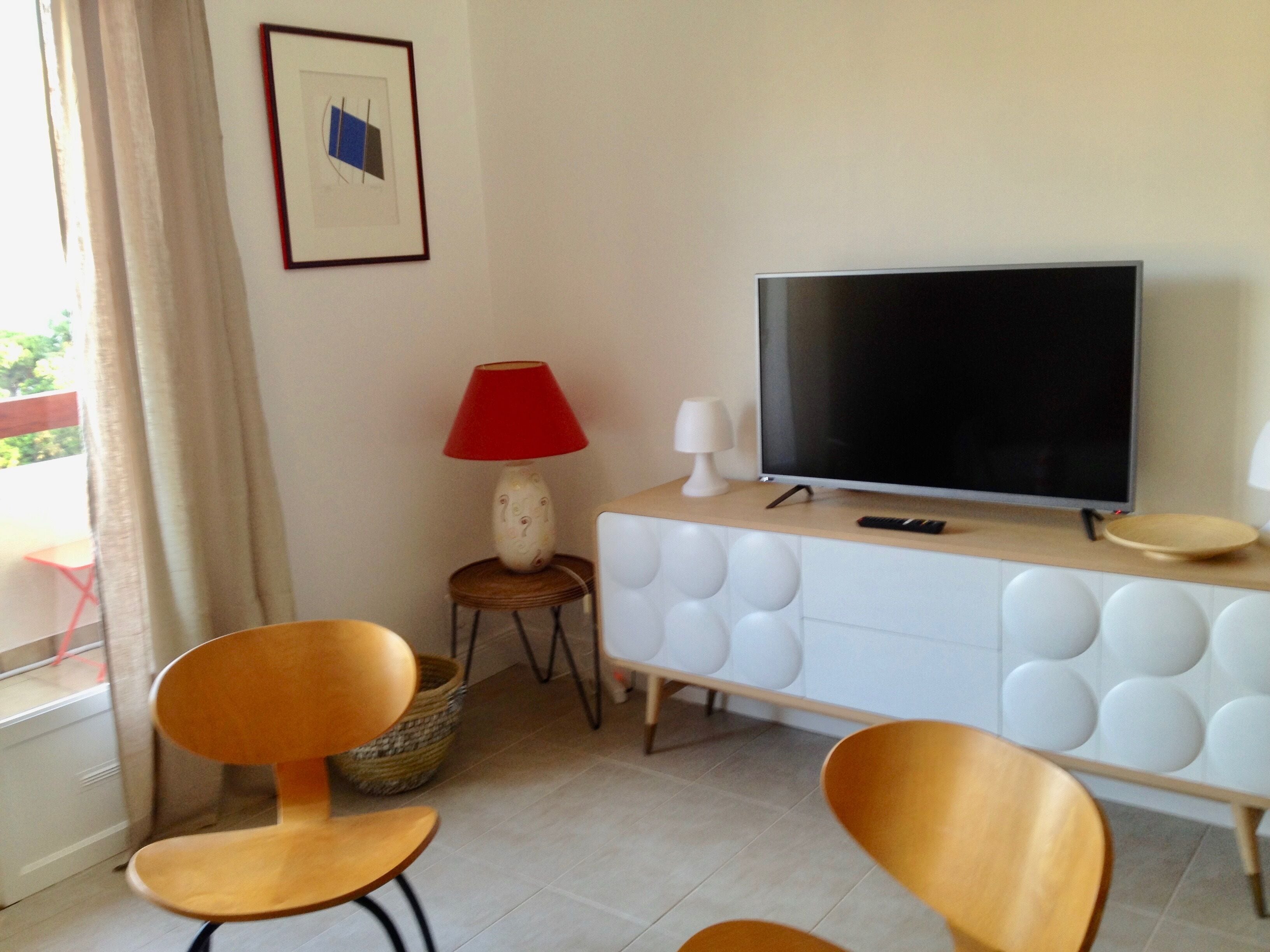 Une chambre moderne équipée d’une télévision, de deux chaises en bois, d’une lampe rouge et d’une armoire blanche.