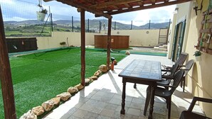 A covered patio with a table and chairs, a view of a green lawn, and a mountainous landscape in the background.