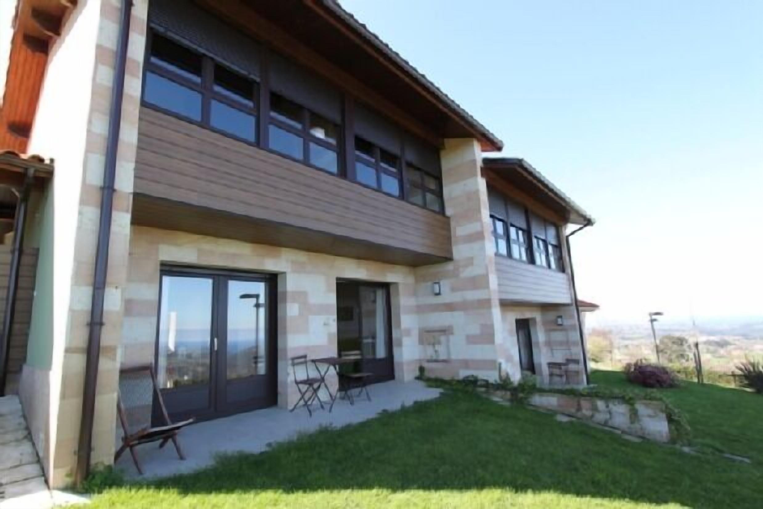 Una casa moderna de dos pisos con amplios ventanales correderos, un patio con mobiliario exterior y vistas al paisaje.