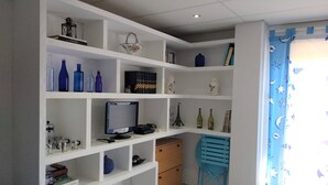 A room with white shelving units containing various items like books, decorative objects, and a computer monitor. There is a small wooden cabinet and a blue chair. - Nevada Apartaments, between Granada and Sierra Nevada (Cenes de la Vega)