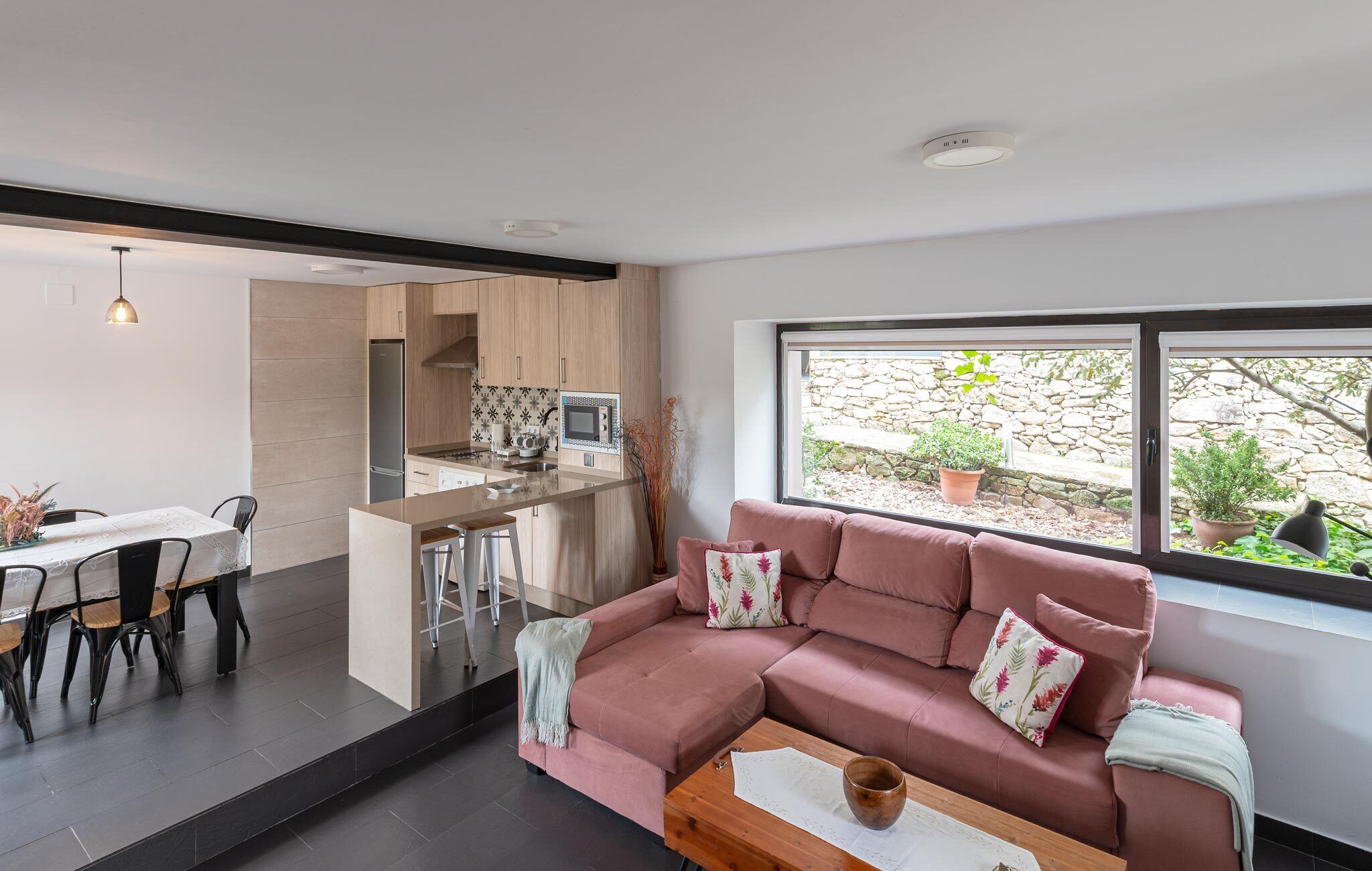 A modern living room with a pink sofa, a dining table with chairs, a kitchen area, and a large window with a view of a stone wall and plants.
