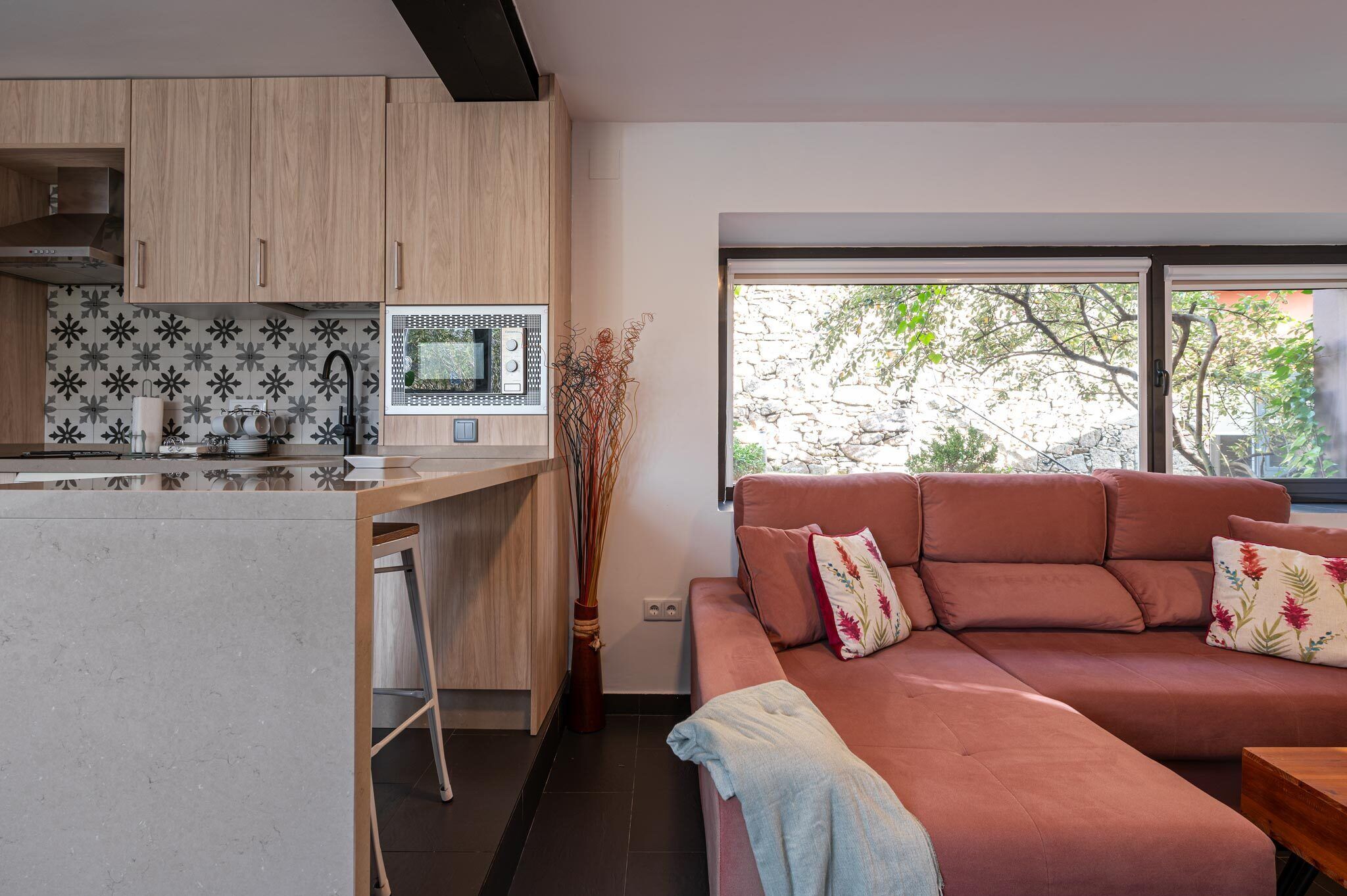 Une cuisine moderne avec des meubles en bois, un four à micro-ondes encastré et un tabouret de bar. Un canapé rouge agrémenté de coussins à motifs floraux se trouve dans l’espace salon, et une grande fenêtre offre une vue sur les arbres.