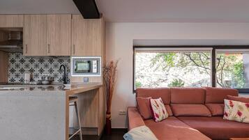 Une cuisine moderne avec des meubles en bois, un four à micro-ondes encastré et un tabouret de bar. Un canapé rouge agrémenté de coussins à motifs floraux se trouve dans l’espace salon, et une grande fenêtre offre une vue sur les arbres.