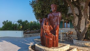 A sculpture of a man and a child in a circular stone base, surrounded by trees and a building with green shutters. - Secluded Villa Dubrava, private pool, olive grove surrounds the villa, Brac Island (Pučišća)