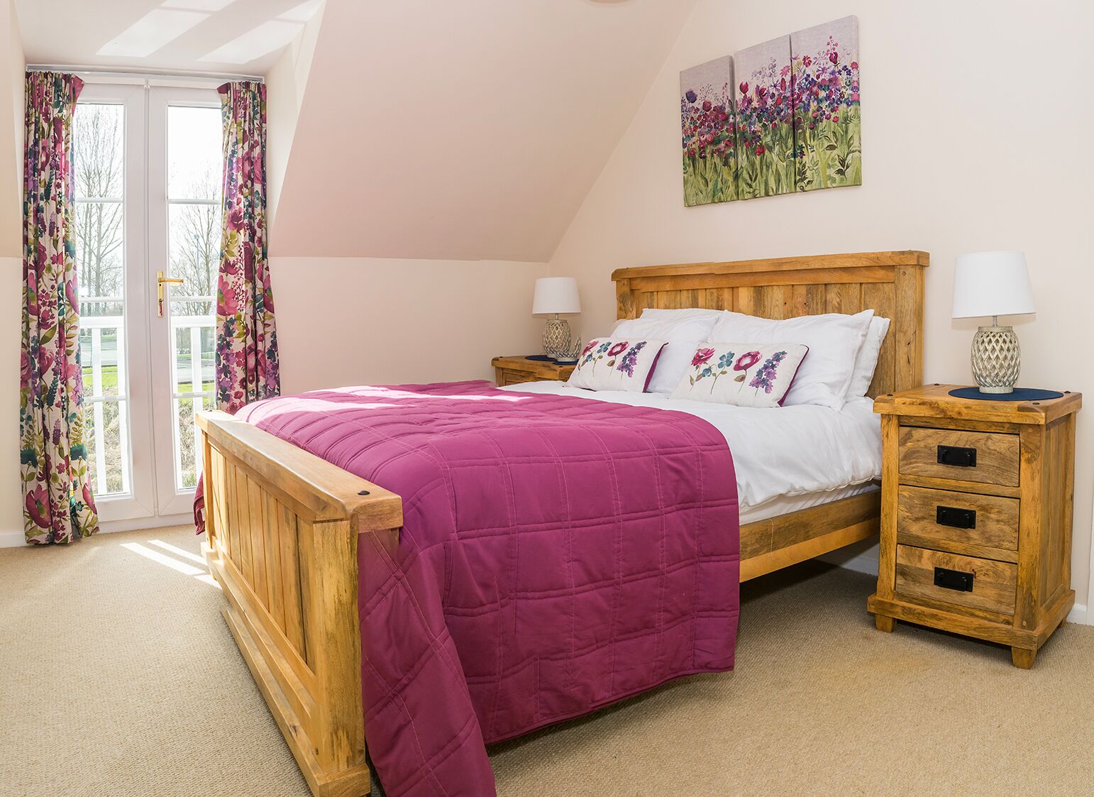 A bedroom with a wooden bed, floral curtains, a painting of flowers, and a nightstand with a lamp.