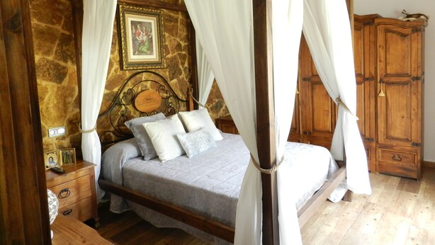 A four-poster bed with white curtains, a stone wall, a wooden cabinet, and a framed picture on the wall.