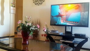 Ein Wohnzimmer mit Fernseher, der einen Film zeigt, einem Glastisch mit Blumen und einem dekorativen Wandbild.