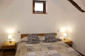 Quarto com cabeceira de madeira, duas mesas de cabeceira com luminárias, colcha estampada com corações e uma pequena janela sobre a cama.