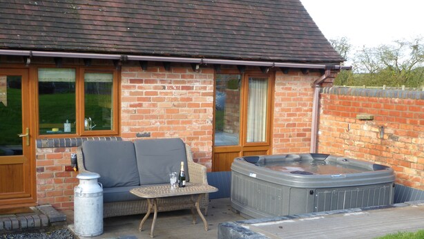 A patio area with a brick wall, a hot tub, a sofa, and a table with a bottle and glass. - Coach House at Oaks Barn Farm, optional salt water hot tub near to Stratford. (Alcester)