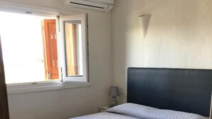 A bedroom with a bed, an air conditioning unit, a window, and a nightstand with a lamp.