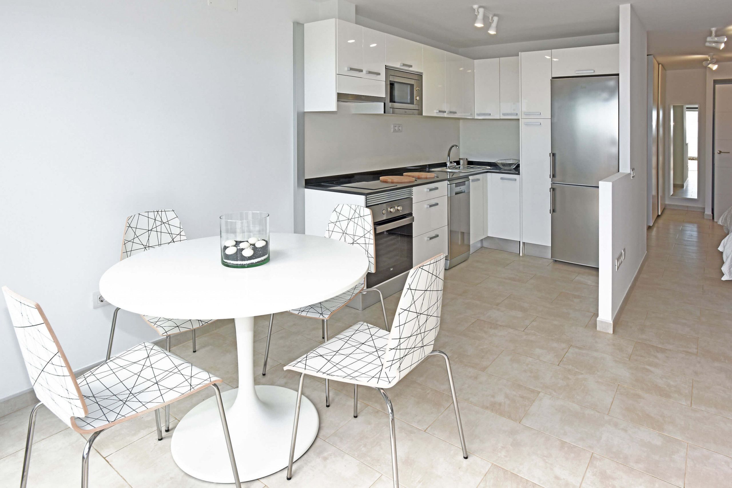 Una cocina moderna con armarios blancos, un refrigerador de acero inoxidable y un comedor con mesa y sillas blancas.
