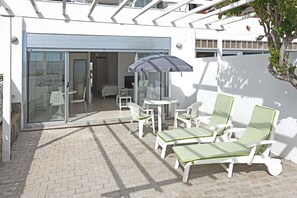 Une terrasse avec des fauteuils de jardin blancs et une table, une porte vitrée donnant sur une pièce, et un arbre.