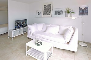 A white living room with a sofa, a coffee table, a TV on a stand, a floor lamp, and a wall with framed pictures. - Chalet Mare Nostrum (Maspalomas)