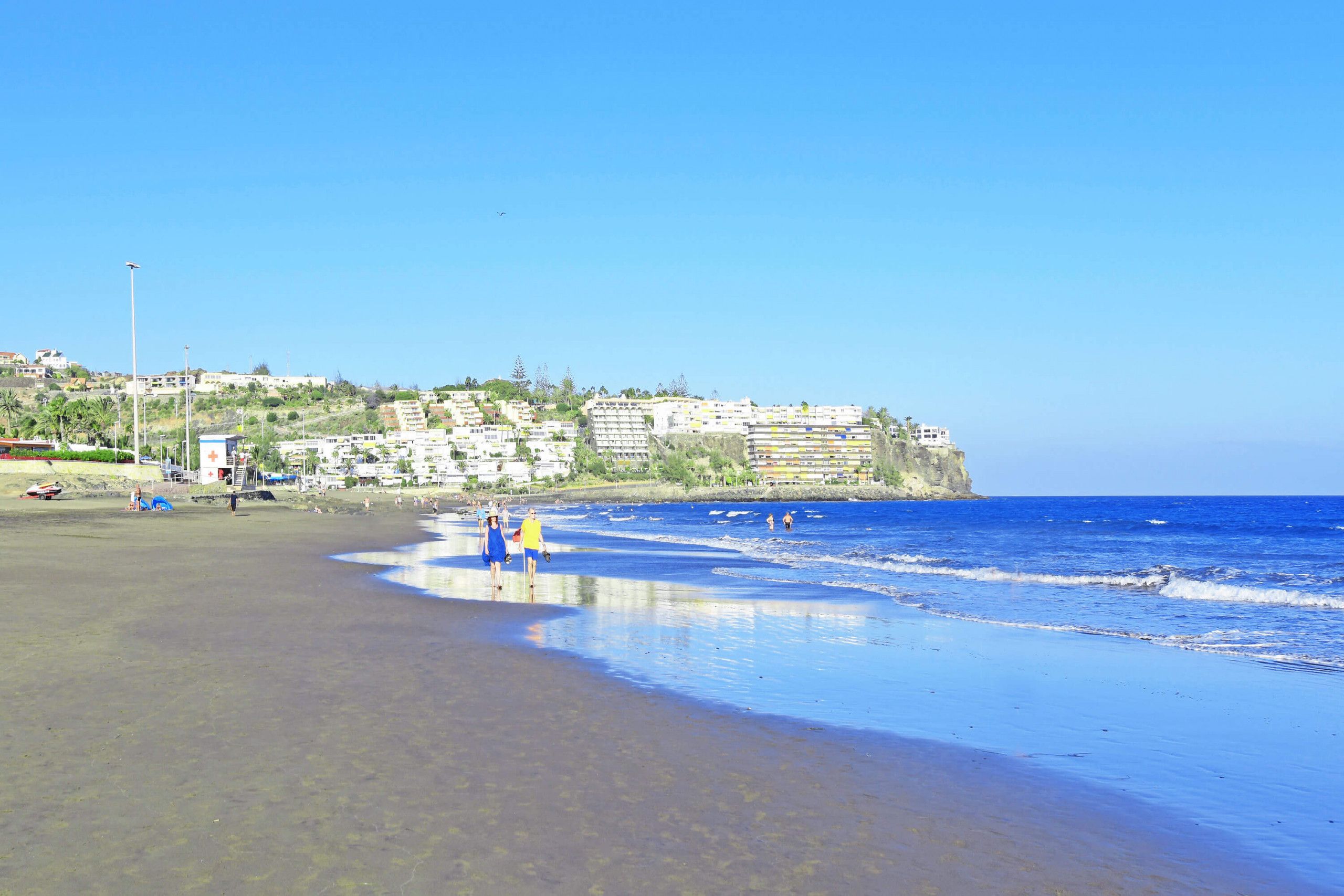 A beach with clear blue water, a few people walking, and buildings in the background.