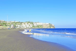 A beach with clear blue water, a few people walking, and buildings in the background. - Chalet Mare Nostrum (Maspalomas)
