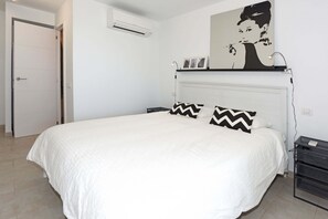 A bedroom with a large bed, a white headboard, a wall-mounted shelf with pictures, and a bedside table with a lamp. - Chalet Mare Nostrum (Maspalomas)