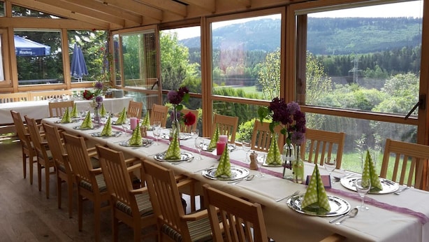 Une longue table à manger, dressée pour un repas, entourée de chaises, avec de grandes fenêtres et une vue sur un paysage boisé.