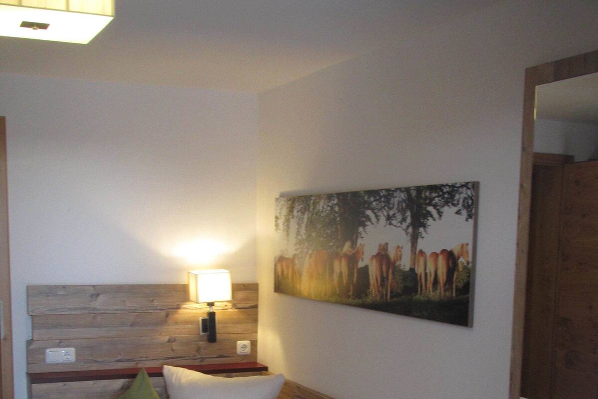 A neatly made bed with a green upholstered headboard, a bedside lamp, and a painting of horses on the wall.