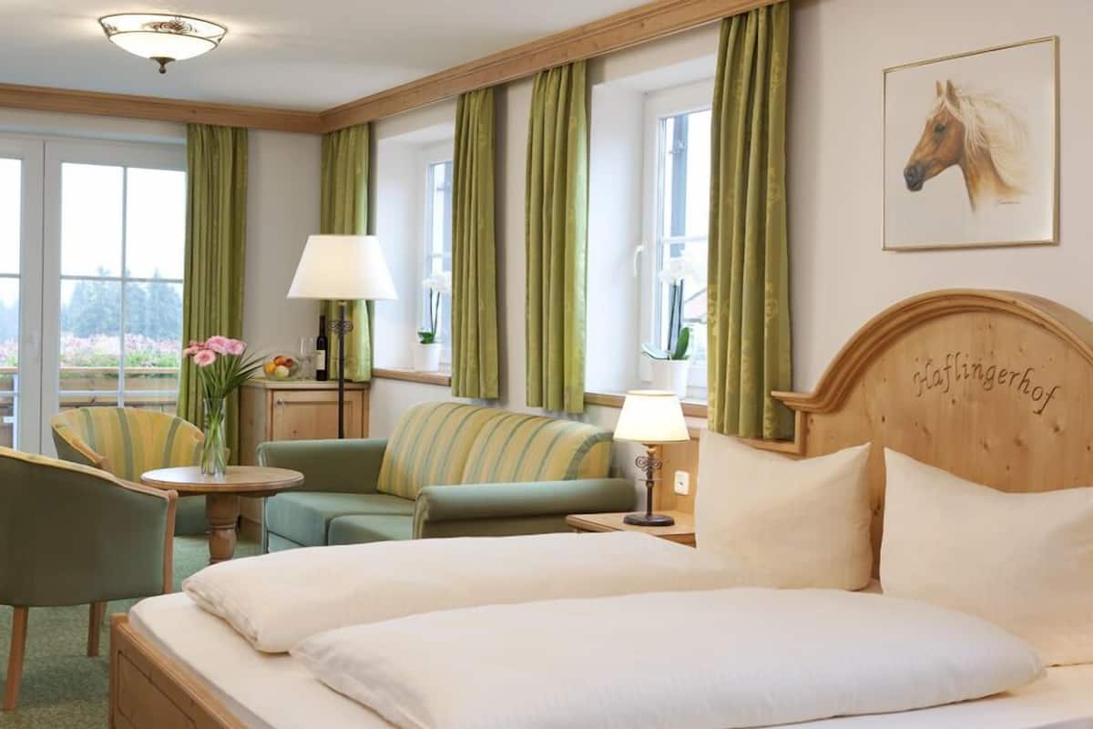 A hotel room with a large bed, two green upholstered chairs, a small table with a vase of flowers, and a lamp.