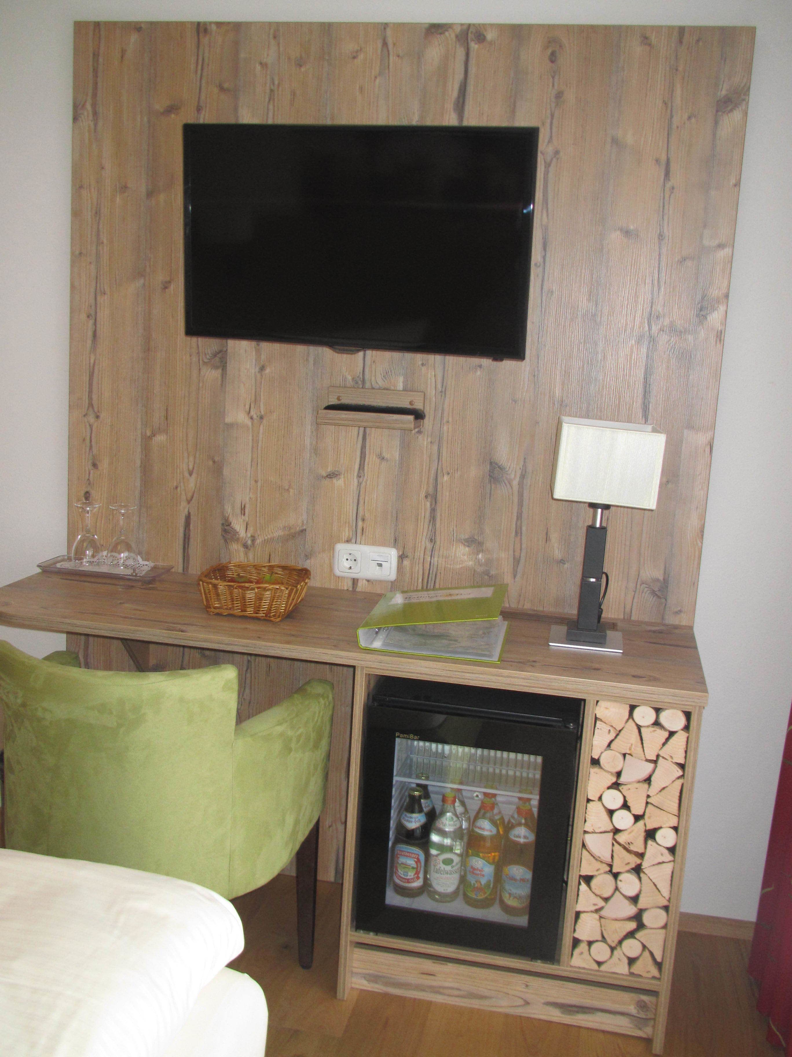 A compact room with a wooden wall-mounted TV, a small refrigerator with beverages, a lamp, and a table with a basket and glasses.