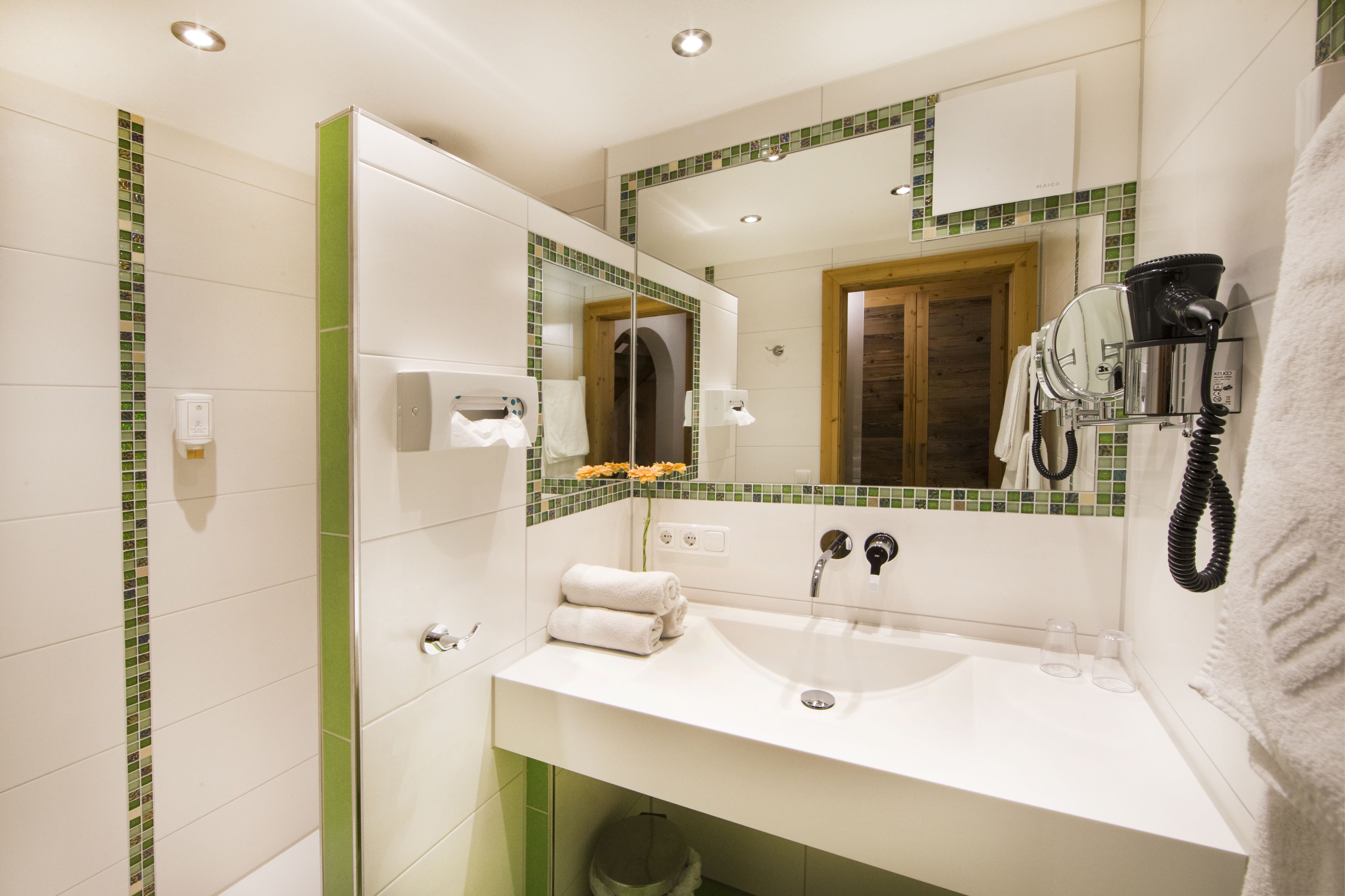 Un baño moderno con un lavabo blanco, salpicadero de azulejos verdes, un espejo grande, un secador de pelo y un perchero para toallas.