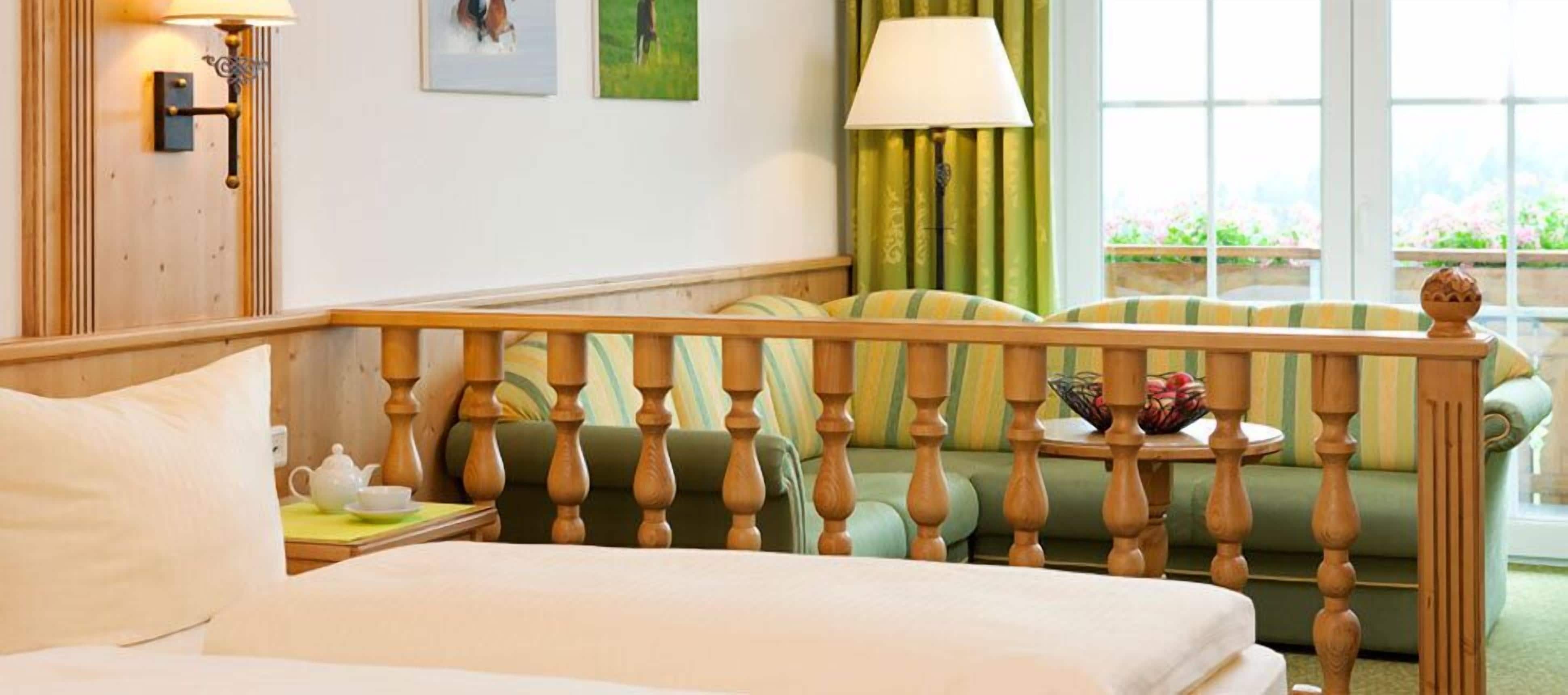 A hotel room with a bed, a wooden headboard, a green sofa, a small table with a bowl of fruit, and a window with curtains.