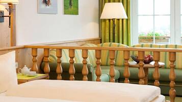 A hotel room with a bed, a wooden headboard, a green sofa, a small table with a bowl of fruit, and a window with curtains.