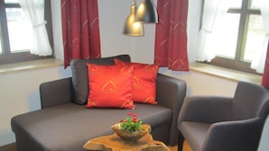 Un salon avec un canapé gris, un coussin rouge, une table basse en bois ornée d’une plante et un fauteuil noir.