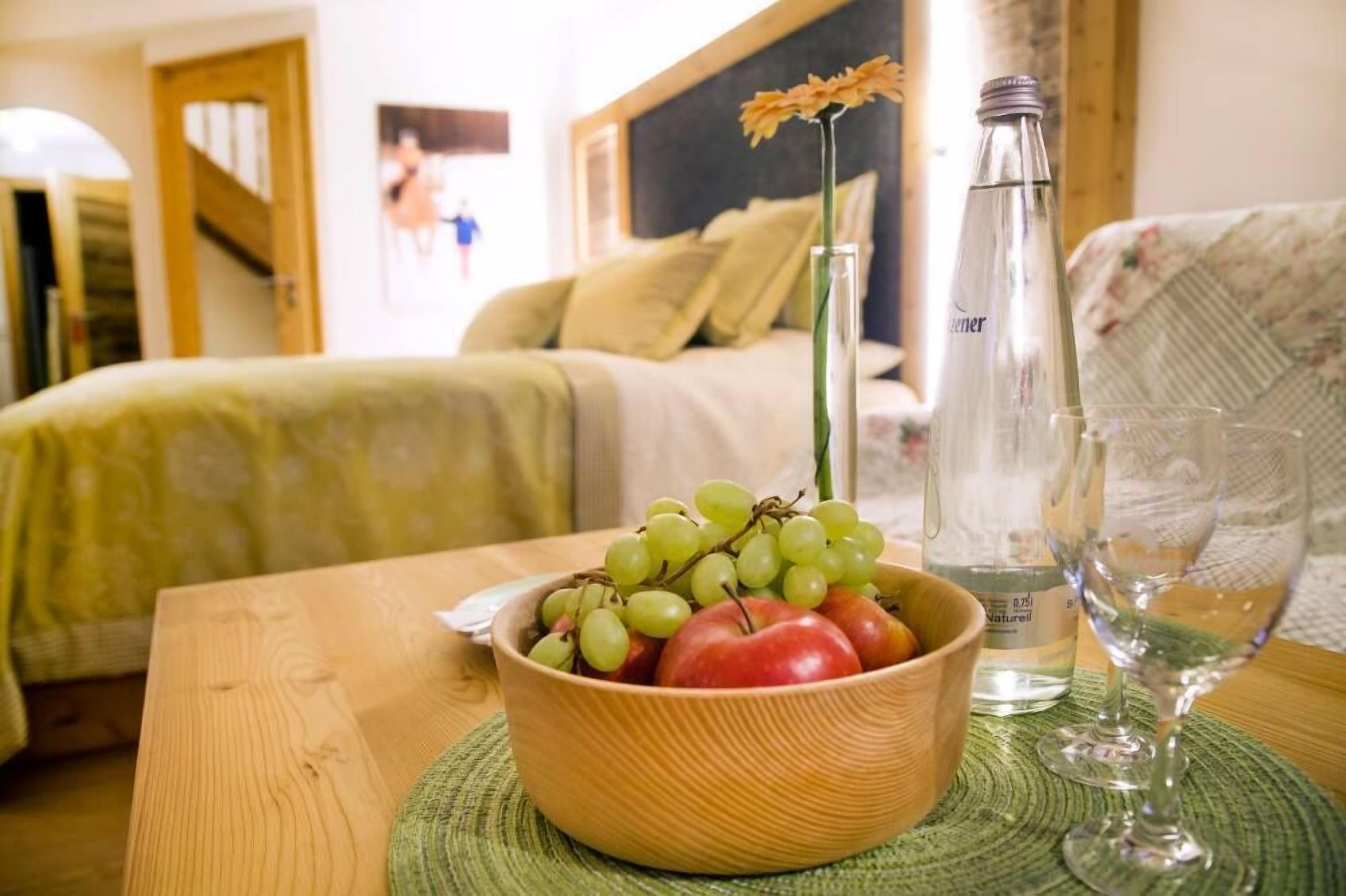 Un saladier en bois contenant des raisins verts et des pommes rouges, posé sur une table, à côté d’une bouteille transparente et d’un verre à vin.