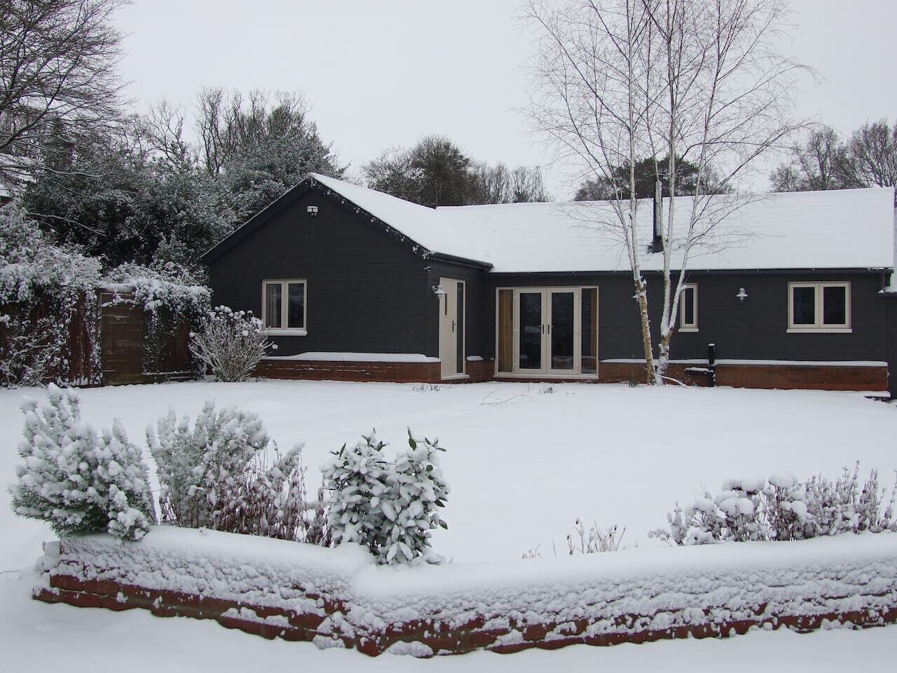 Une maison aux couleurs sombres, avec un toit et une cour couverts de neige.