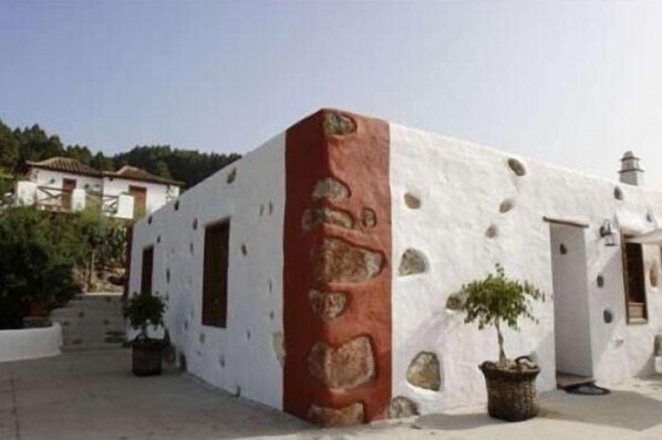 A white building with a red column and stone details, surrounded by potted plants and a clear sky.